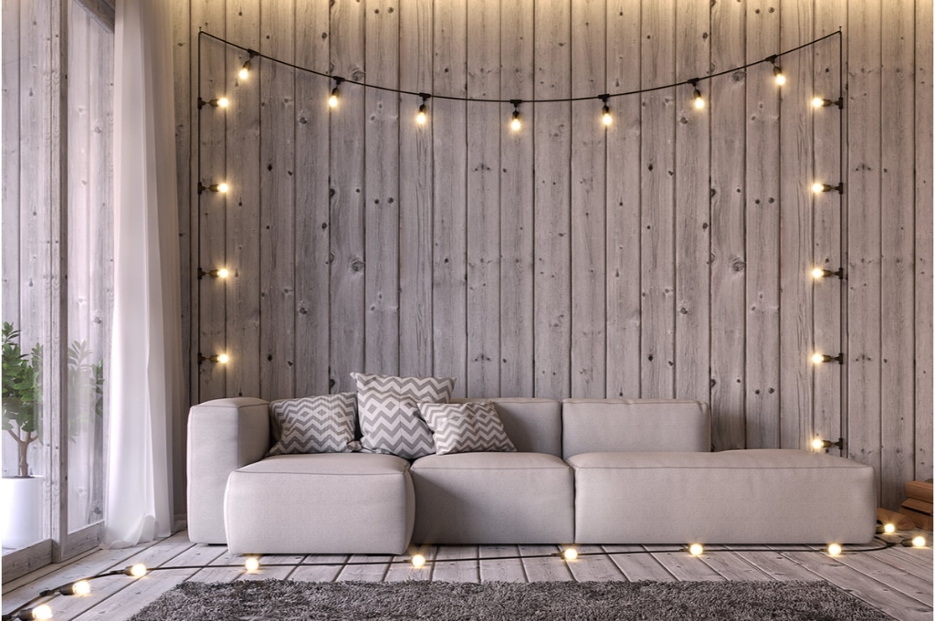 dans un salon entièrement en bois gris clair du sol aux murs, une guirlande lumineuse est allumée et accrochée au-dessus d'un canapé d'angle gris. 3 coussins sont disposés sur le canapé d'angle.