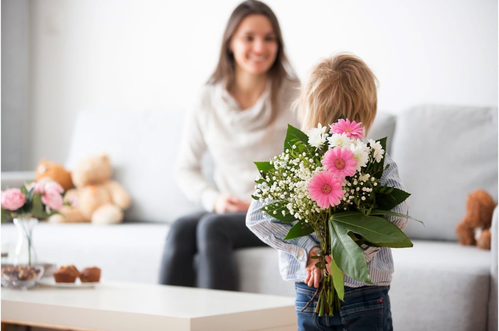 un petit garçon tient un bouquet de fleurs dans son dos. Il est dans le salon devant sa maman assise sur le canapé. Elle va recevoir la surprise d'un beau bouquet de fleurs. Des ours en peluche sont posés sur le canapé à côté d'elle.
