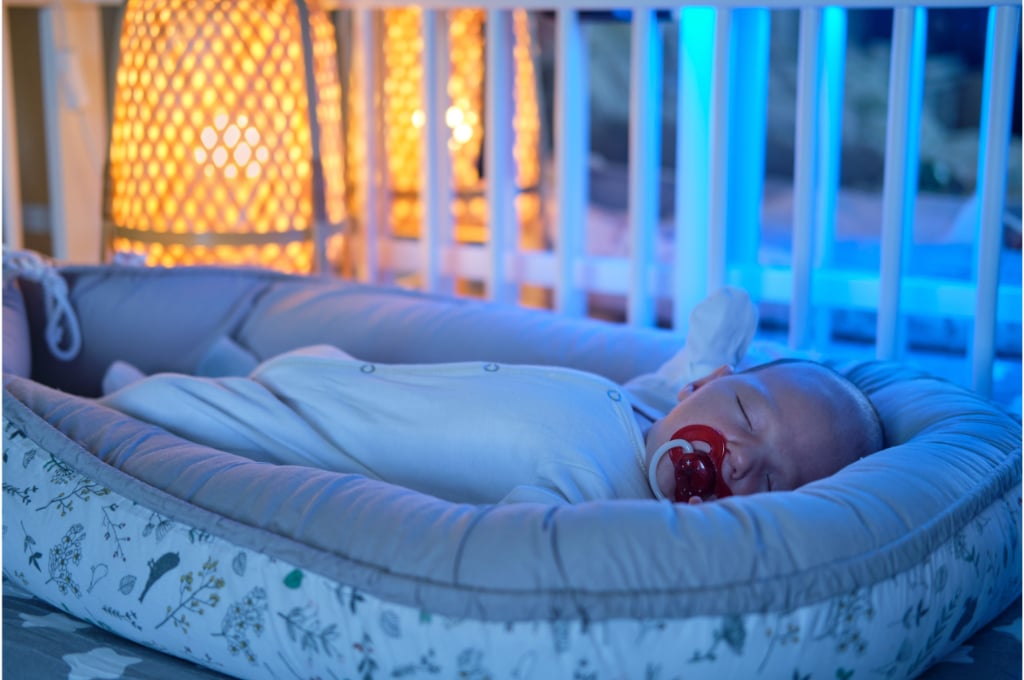 dans un berceau à peine éclairé, un bébé dort entouré de son réducteur de lit. il a une tétine rouge à la bouche et est en pyjama.