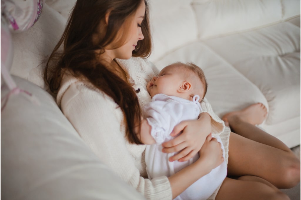 sur un canapé beige, une maman est posée avec son bébé endormi dans les bras. elle le regarde tendrement. son bébé porte une robe blanche et est endormie contre le torse de sa maman.