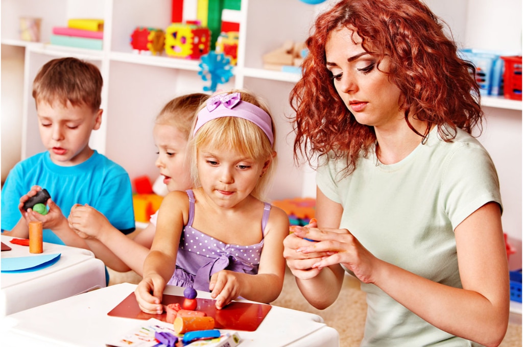 une femme rousse assise à une table avec d'autres enfants, manipule avec eux de la pâte à modeler. Divers jouets sont disposés derrière eux sur des étagères.