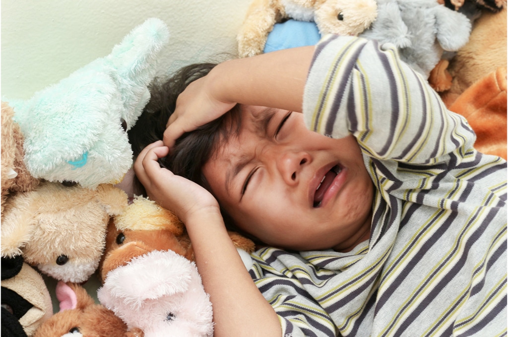 vue rapprochée sur un enfant qui pleure et se tient la tête au milieu de peluches. les manches de son haut à rayures sont retroussées. 