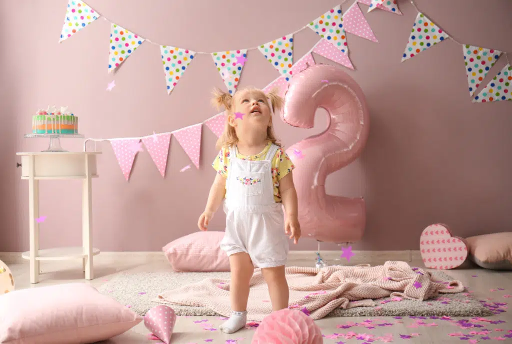 chambre rose de petite fille, décorée pour son anniversaire avec des banderoles et un énorme ballon rose en forme de chiffre 2. une petite fille au centre regarde vers le plafond.