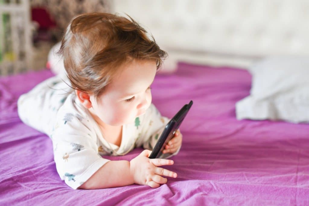 un bébé couché sur le ventre sur un lit regarde l'écran d'un téléphone mobile. il porte un body blanc avec des dessins colorés.