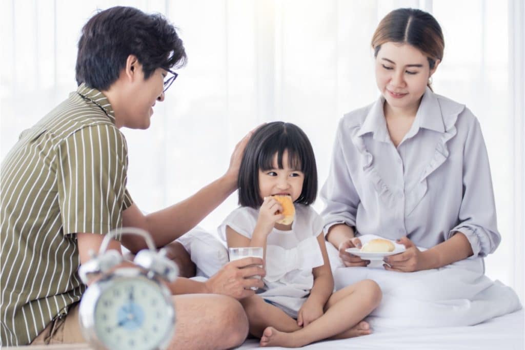 une famille d'origine asiatique est assise en tailleur. Le père en chemise à rayures, met une main sur la tête de sa petite fille qui mange une tartine. La mère à côté tient une assiette avec une brioche dedans.