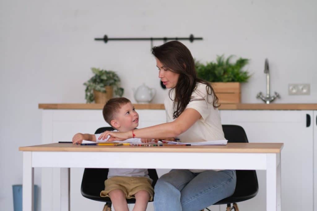 dans une cuisine, une femme est assise à une table avec son enfant garçon. elle touche des papiers avec des crayons de couleurs sur la table et son garçon la regarde.