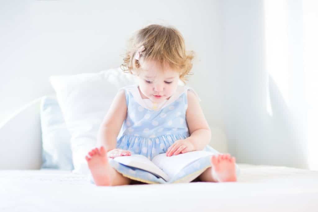 une petite fille est assise sur un grand lit et lit un livre posé sur ses jambes. elle porte une robe bleue à pois blancs.