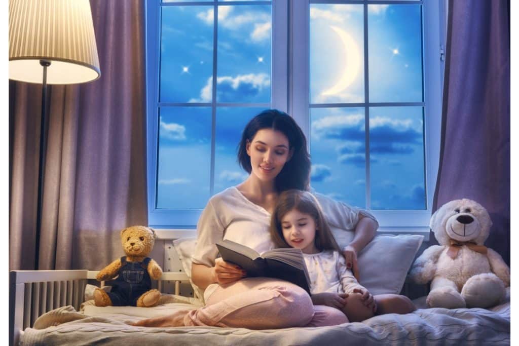 dans un lit d'enfant, une maman lit une histoire à sa petite fille devant une fenêtre avec un grand croissant de lune.