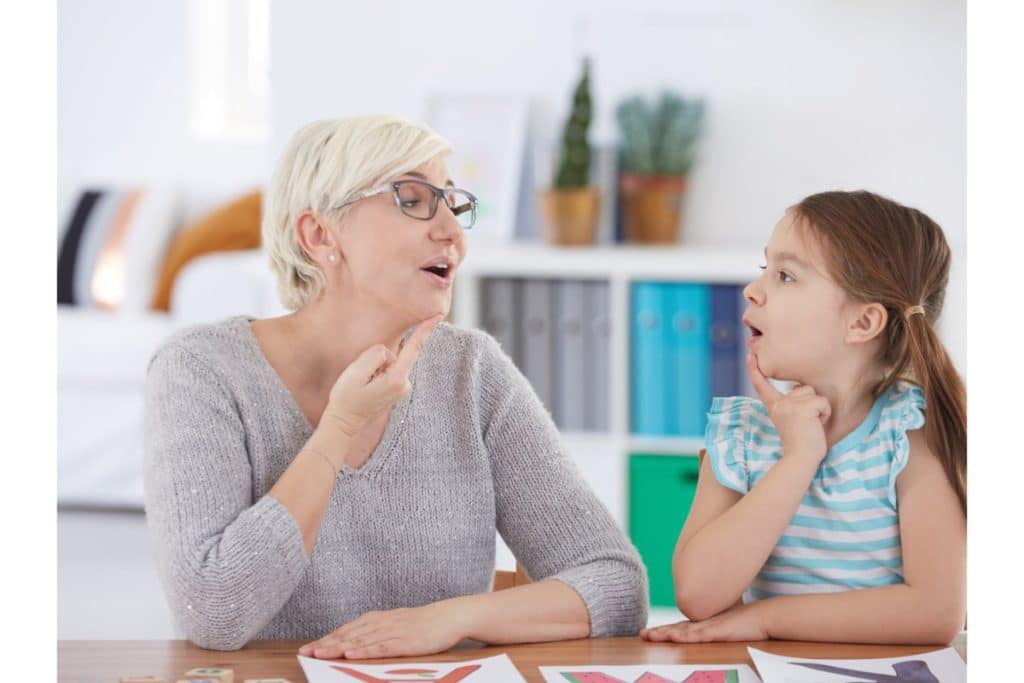 une orthophoniste assise à une table avec une jeune fille, met son doigt sur le menton en ouvrant la bouche. La jeune fille imite ses gestes en la regardant.