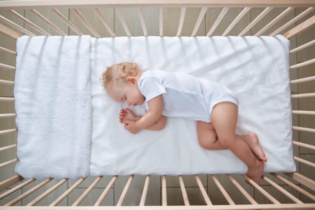 vue du dessus d'un bébé endormi dans un lit à barreaux sur un matelas. il porte un body blanc à manches courtes.