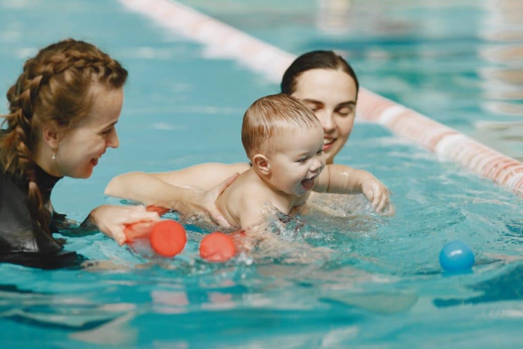 2 femmes nagent une piscine municipale avec un bébé nu qui barbote dans l'eau.