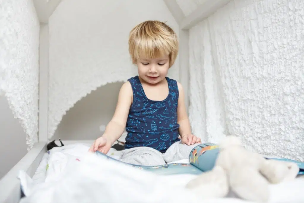 un enfant blond est assis dans son lit cabane avec un livre ouvert qu'il regarde