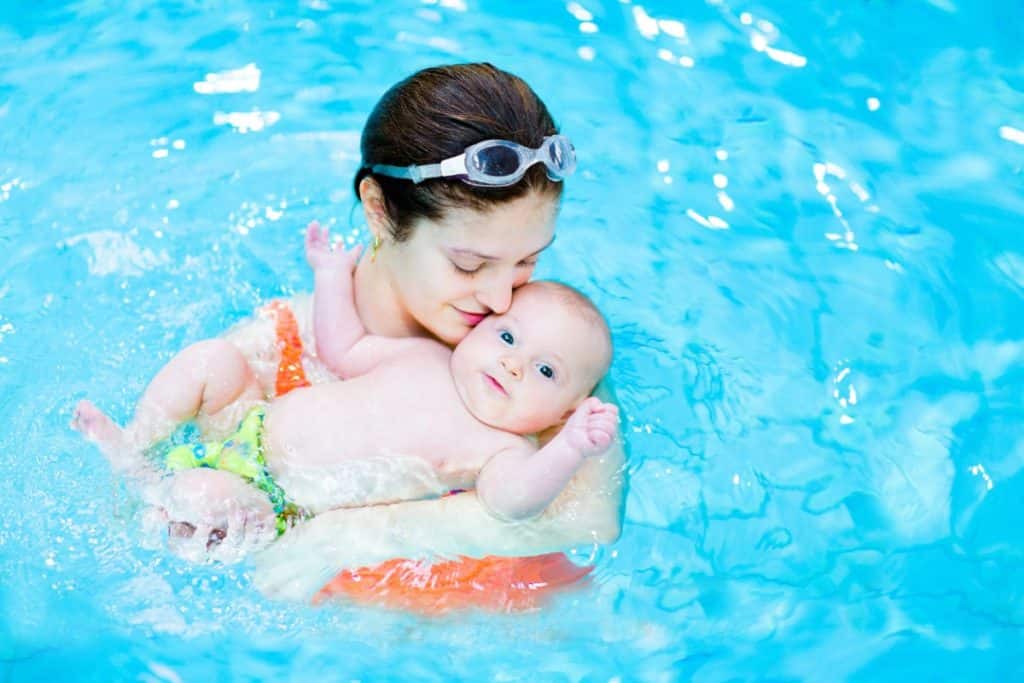 dans une piscine, une femme tient son bébé entre les bras qui est sur le dos en train de nager.