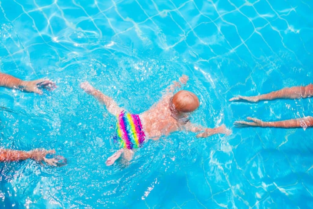 dans une piscine vue du dessus, un bébé nage la tête sous l'eau vers des bras tendus devant lui. d'autres bras sont tendus derrière lui.