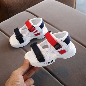 Paire de sandale blanche ouverte pour bébé avec une bande scratch bleue et une autre rouge, montrée par une main pour une chaussure et l'autre est posée en arrière plan sur un support gris