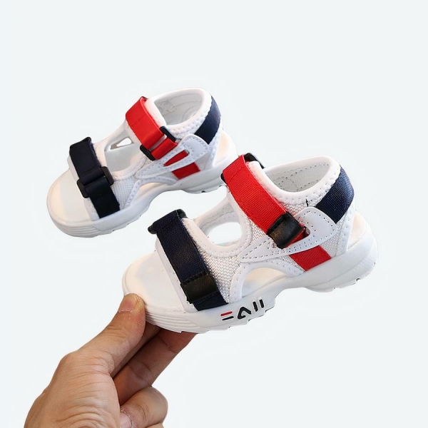 Paire de sandale blanche ouverte pour bébé avec une bande scratch bleue et une autre rouge, montrée par une main pour une chaussure et l'autre est posée en arrière plan sur un support gris