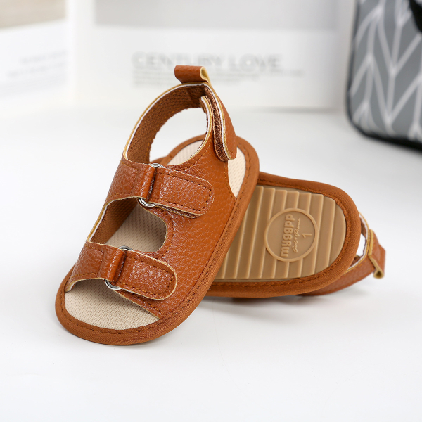 Sandale marron plate d'été pour bébé ouverte, avec 2 bandes de scratch sur le dessus posée l'une sur l'autre pour les présenter, sur un sol blanc et brillant