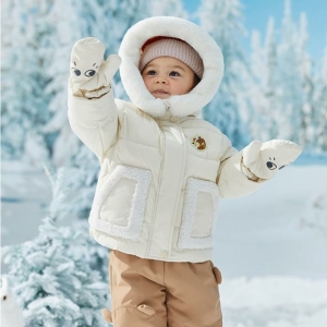 enfant en train de jouer dans la neige, emmitouflé dans une doudoune pour bébé beige avec une grosse capuche douillette , et des moufles assorties